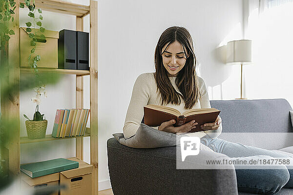 Lächelnde Frau  die auf dem Sofa im Wohnzimmer ein Buch liest