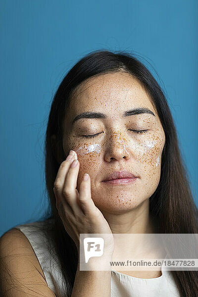 Woman with eyes closed applying moisturizer against blue background
