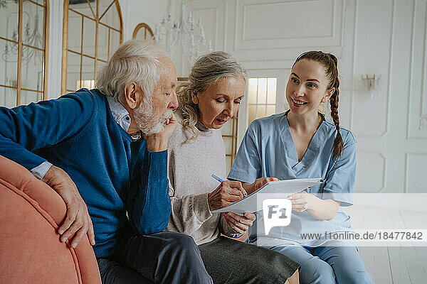 Senior woman signing document with man and caregiver at home