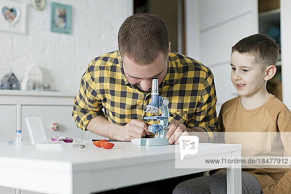 Smiling boy sitting by father looking through microscope at table