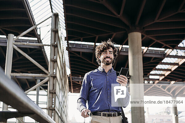 Smiling businessman with smart phone standing at station