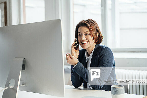 Happy businesswoman talking on smart phone in front of desktop at workplace