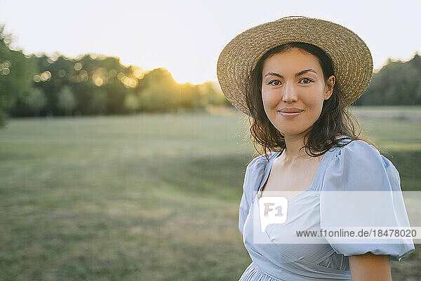 Lächelnde Frau mit Hut steht bei Sonnenuntergang im Park