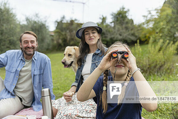 Girl looking through binoculars with parents sitting in background at family picnic