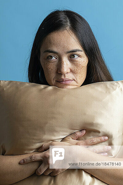Thoughtful woman resting chin on silk pillow against blue background