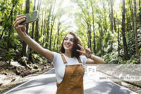 Happy young woman showing peace sign gesture and taking selfie in front of trees