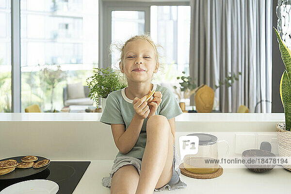 Smiling girl eating pancake sitting on kitchen counter at home
