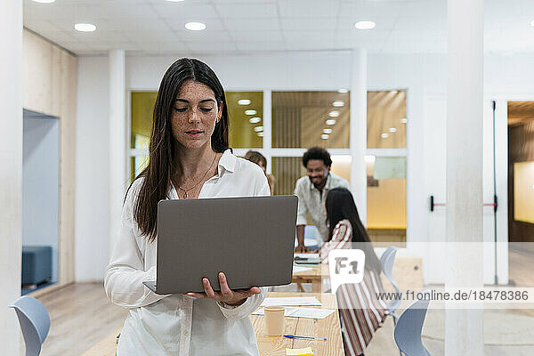 Businesswoman using laptop in office with colleagues in background
