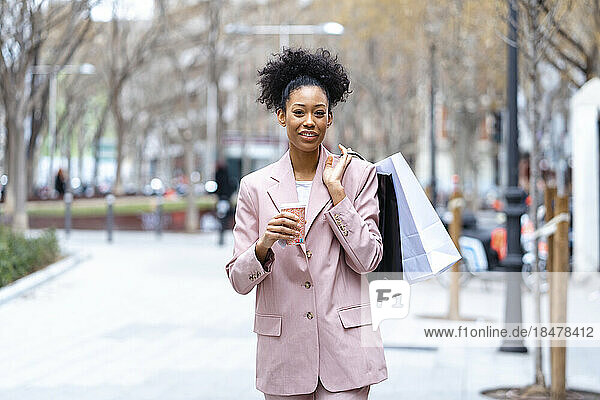 Happy businesswoman with coffee cup and shopping bag standing at footpath