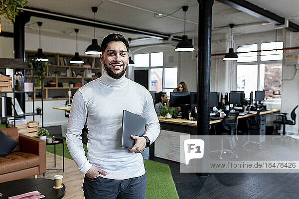Smiling young businessman standing with laptop in office