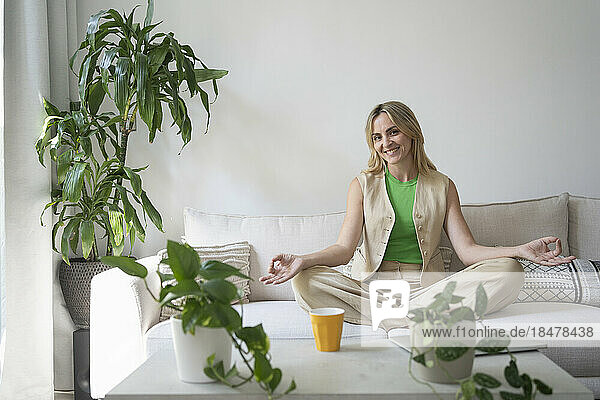 Smiling female freelancer meditating in lotus position on sofa at home