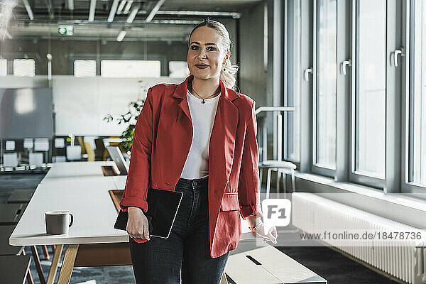 Smiling businesswoman wearing red blazer standing near desk at office
