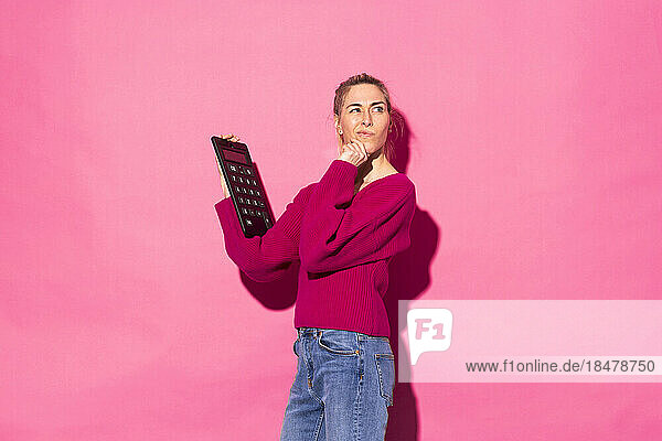 Confused woman with calculator standing against pink background