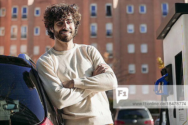 Lächelnder junger Mann steht an einem sonnigen Tag neben einem Elektroauto