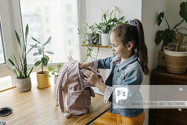Smiling girl opening backpack on table at home