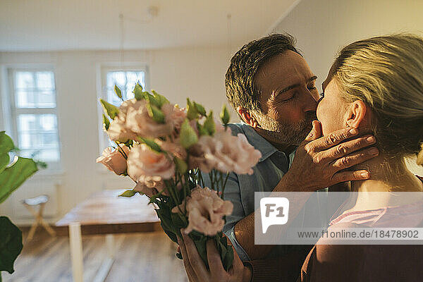 Affectionate husband kissing wife holding bunch of flowers at home
