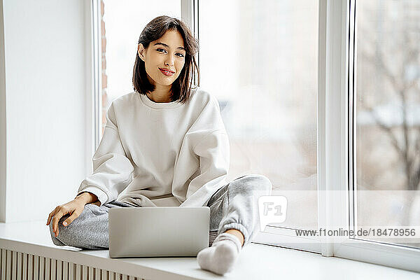 Glückliche Frau sitzt mit Laptop auf der Fensterbank