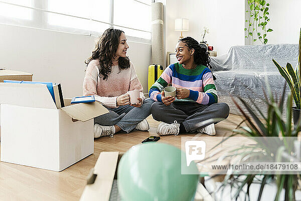 Happy young friends sitting with coffee cup on floor at home