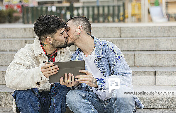 Romantic gay couple kissing with tablet PC sitting on steps