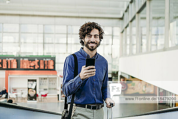 Lächelnder Geschäftsmann mit Mobiltelefon steht am Bahnhof