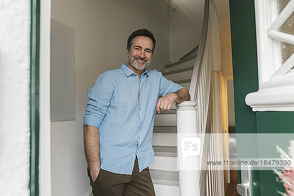 Smiling mature man with hand in pocket standing near staircase at home