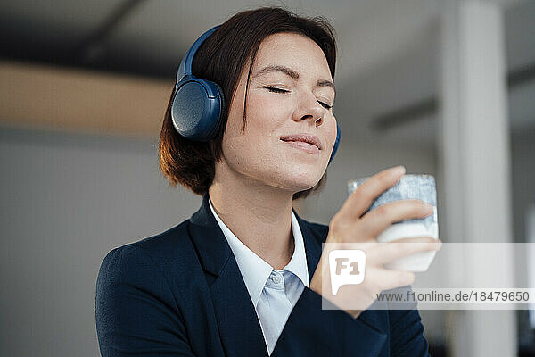 Young businesswoman wearing headphones listening to music holding coffee cup in office