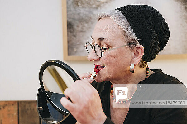 Senior woman applying red lipstick looking in mirror at home