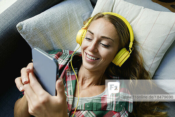Happy woman using mobile phone wearing headphones lying on couch at home