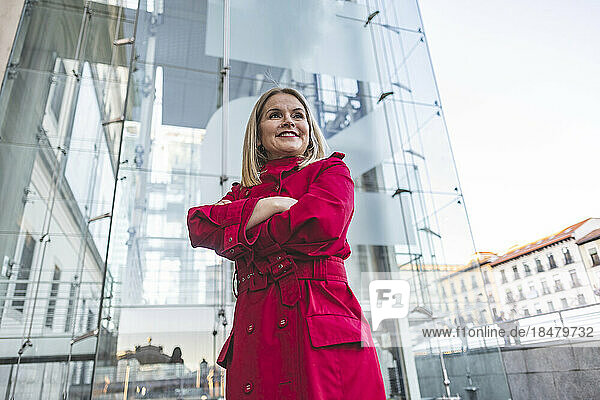 Smiling mature woman with arms crossed standing in front of modern glass building