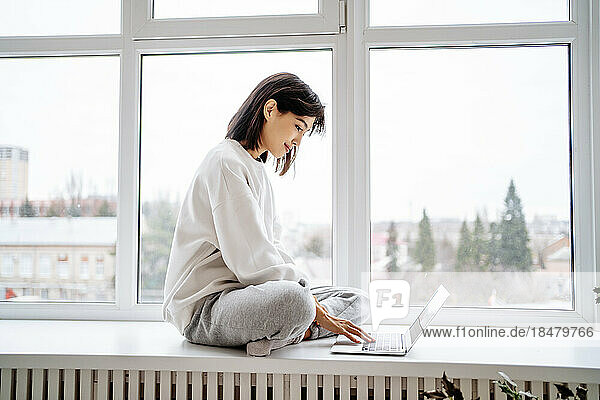 Smiling woman using laptop on window sill at home