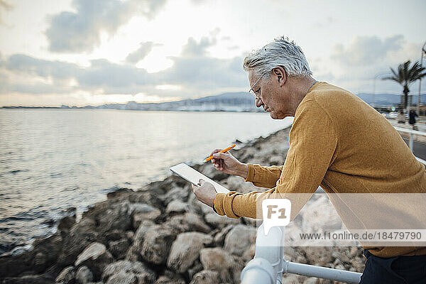 Senior man using tablet PC leaning on railing in front of sea