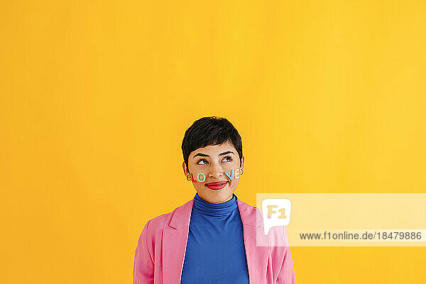 Thoughtful young woman with Love text stickers on face against yellow background
