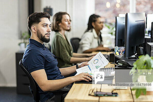 Junger Geschäftsmann mit Diagrammen  der am Desktop-PC im Büro arbeitet