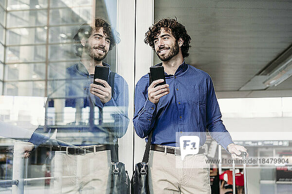 Lächelnder junger Geschäftsmann mit Smartphone  der durch das Fenster schaut