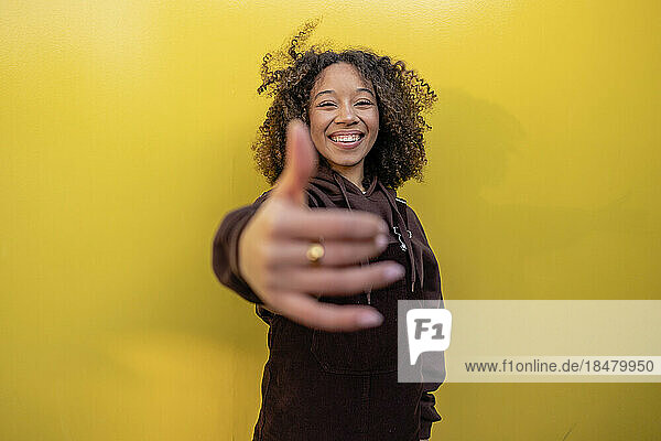 Happy woman with curly hair gesturing in front of yellow wall