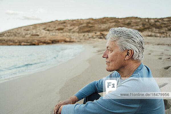 Thoughtful senior man sitting on beach at sunset