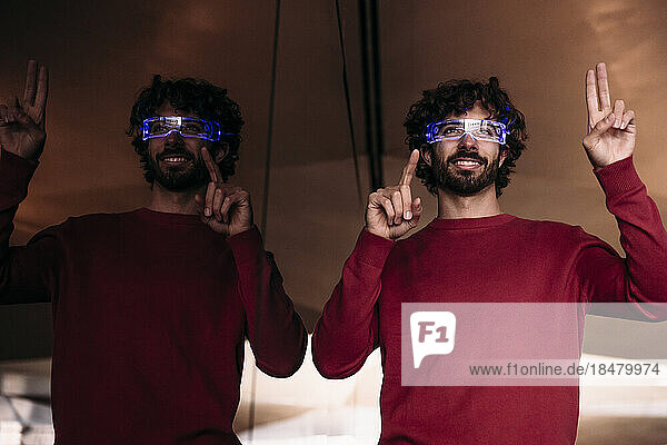 Happy man wearing smart eyeglasses gesturing by glass wall