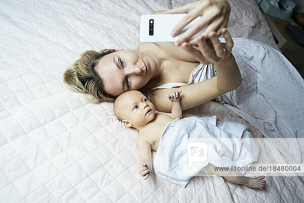 Mother taking selfie with baby boy through smart phone on bed at home