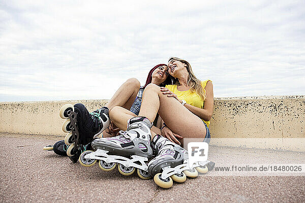 Happy women wearing inline skates sitting at promenade in front of cloudy sky