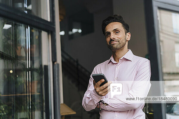 Smiling thoughtful mature businessman with smart phone leaning by door