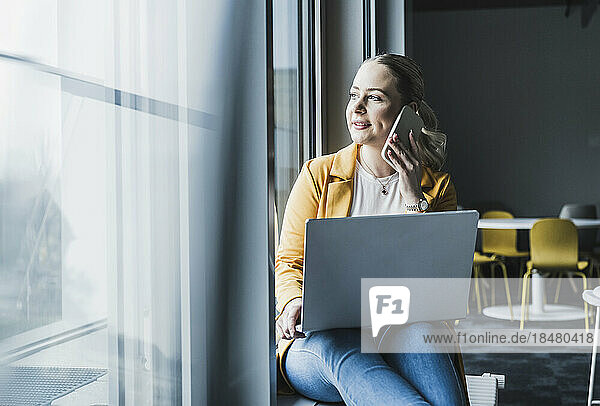 Lächelnde Geschäftsfrau  die am Fenster mit Laptop im Büro sitzt und am Smartphone spricht