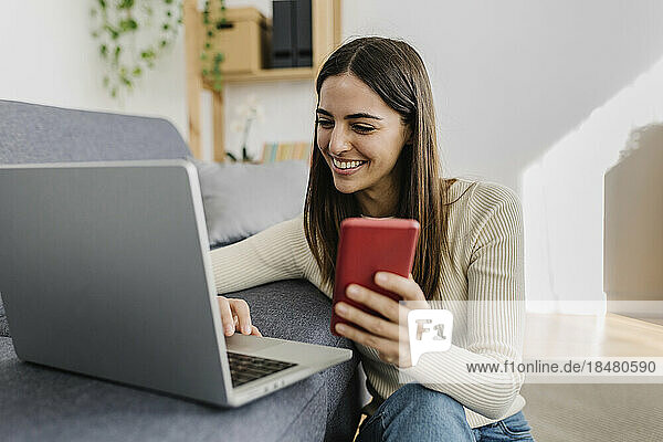 Happy young woman using laptop with smart phone at home