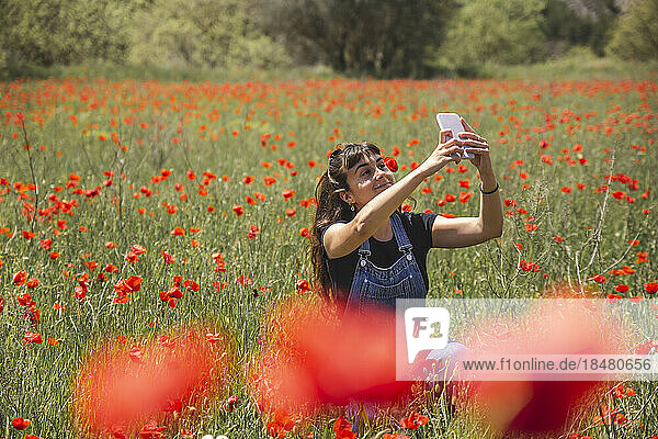 Smiling young woman taking selfie through smart phone amidst field of poppies