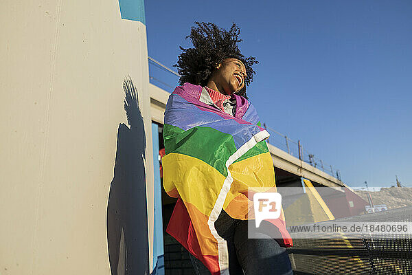 Fröhliche junge Frau  gehüllt in eine Regenbogenfahne  genießt einen sonnigen Tag