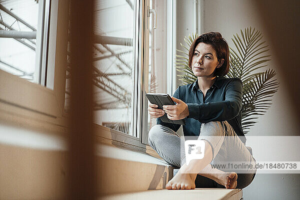 Nachdenkliche junge Geschäftsfrau mit Tablet-PC sitzt auf der Fensterbank im Büro
