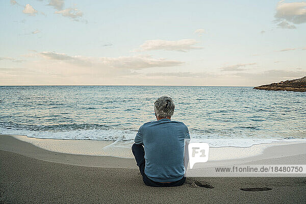 Älterer Mann sitzt auf Sand vor dem Meer am Strand