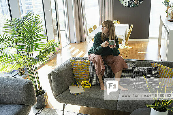 Thoughtful woman with tea cup sitting on sofa at home