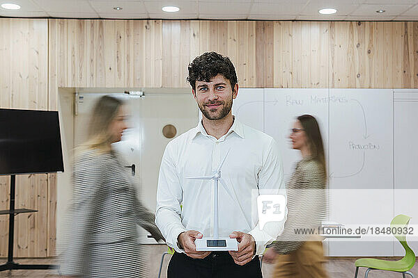 Businessman in office holding wind turbine model with businesswomen passing by
