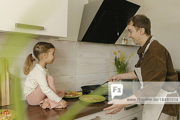 Vater bereitet Pfannkuchen für Tochter zu  die zu Hause auf der Küchentheke sitzt
