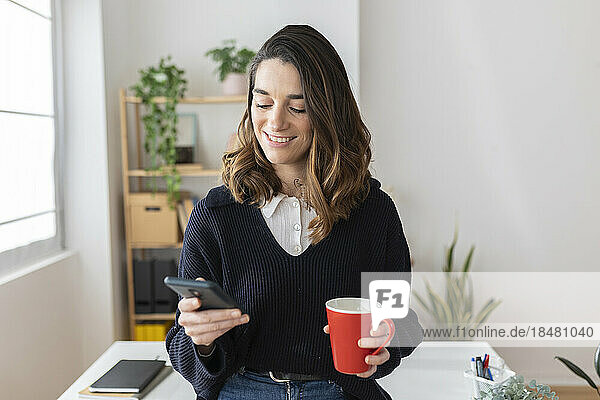 Lächelnde Geschäftsfrau mit Kaffeetasse und Smartphone im Büro